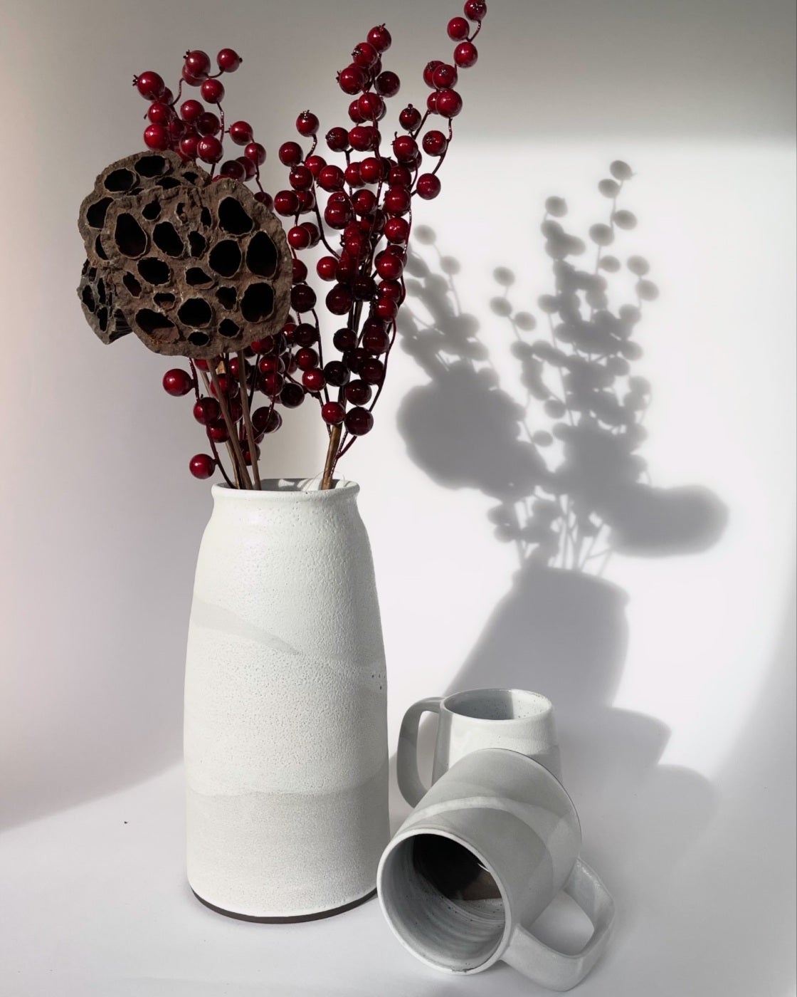 White bud vase holding dried lotus pods and red berries, with a small grey cup