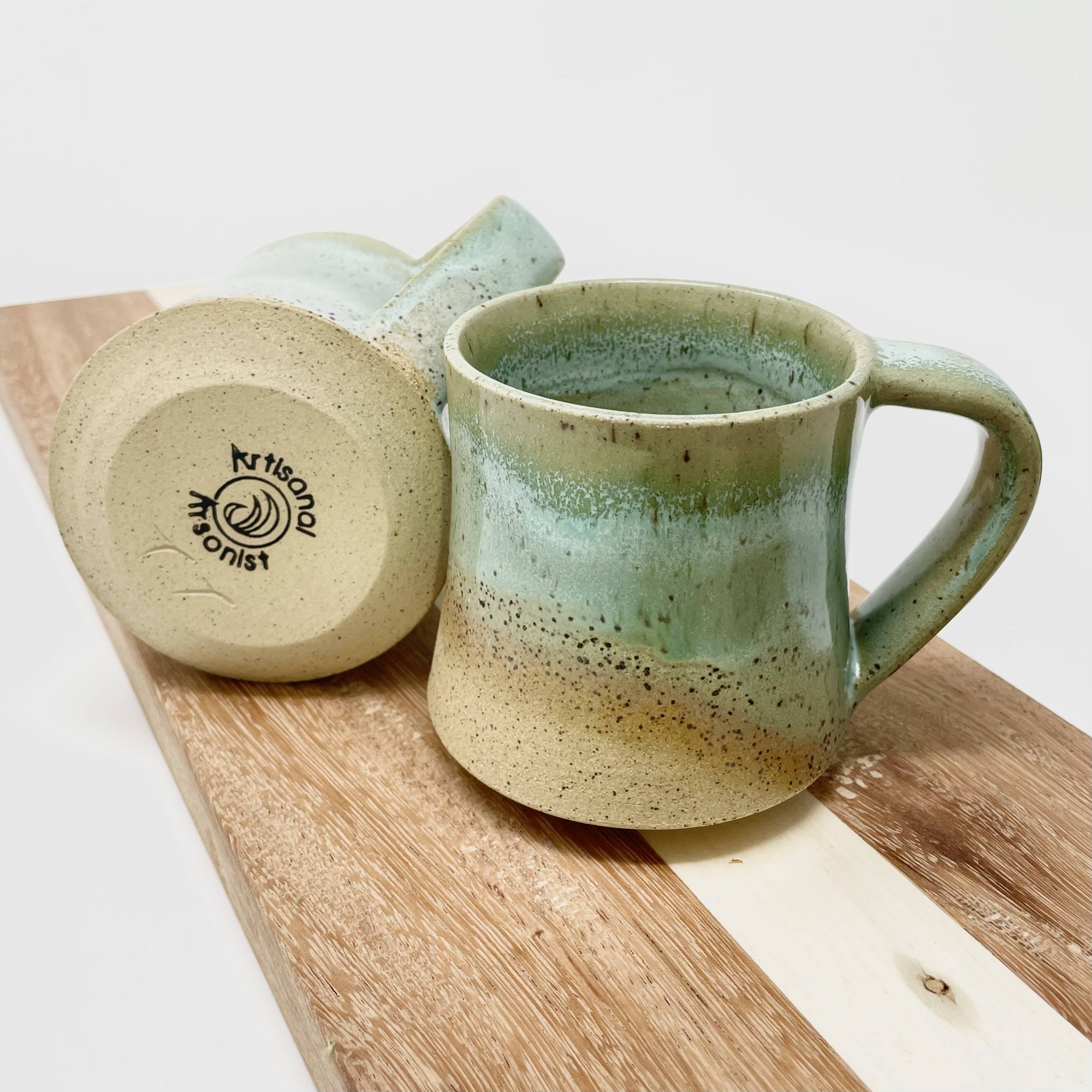 Two speckled cream-and-green glazed mugs on a wooden board, one showing the maker's stamp
