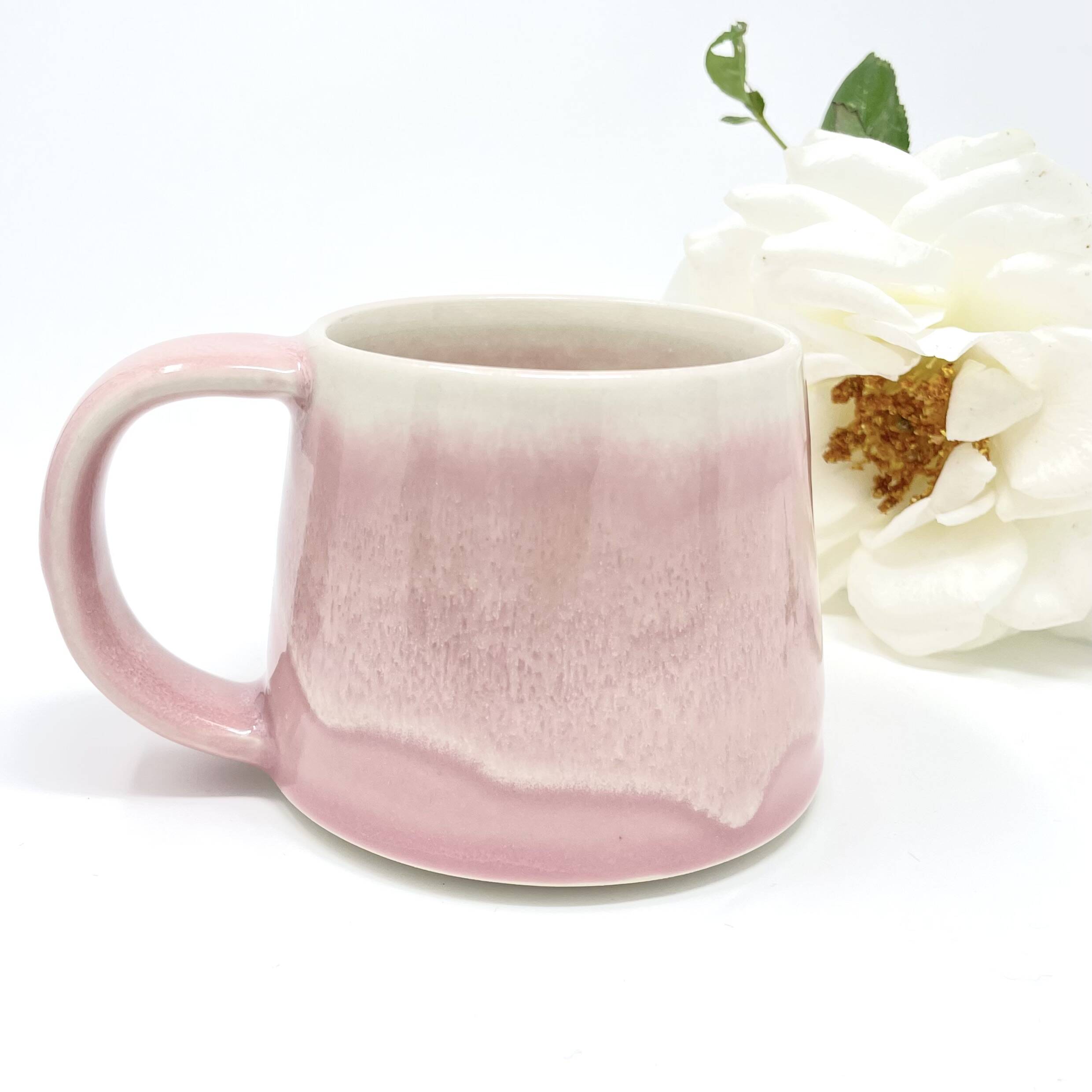 Handmade mug with a soft pink drip glaze beside a white flower