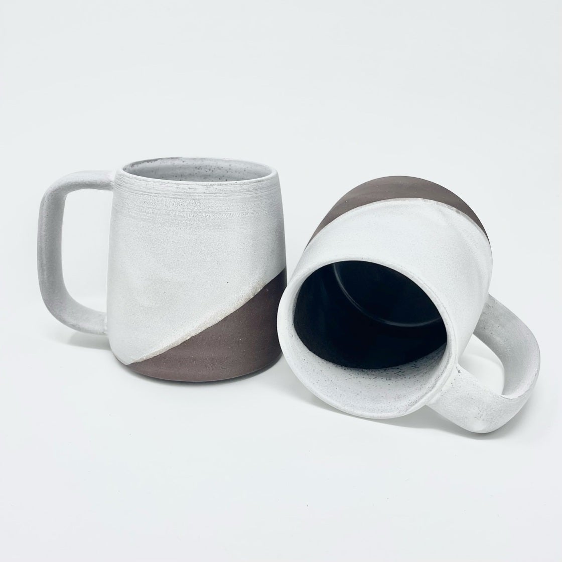 Two mugs with white glaze over dark clay, one on its side revealing a dark blue interior