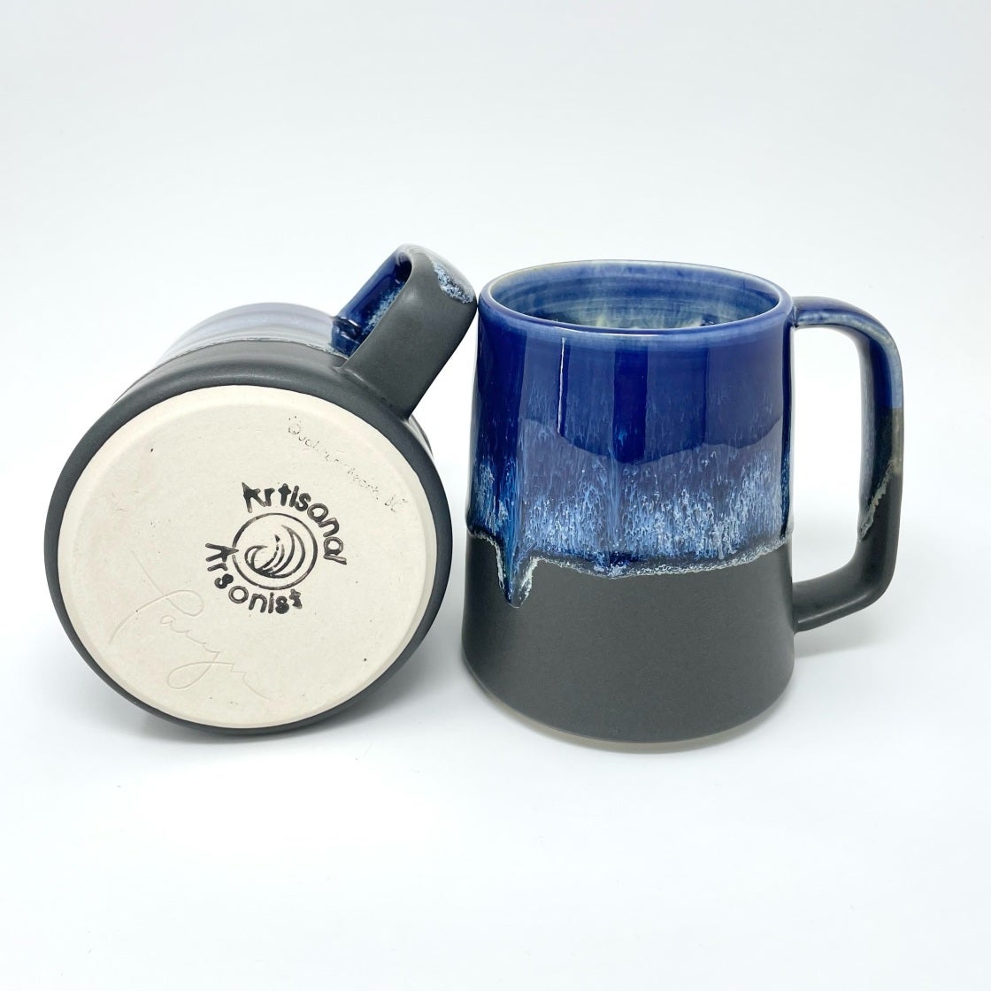Two mugs with a cobalt-blue drip glaze over grey, one showing the maker's stamp